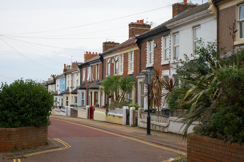 A row of terraced UK homes with brick facades and bay windows, each with small front gardens bordered by low brick walls and black metal fencing. Some gardens feature neatly trimmed bushes and small p